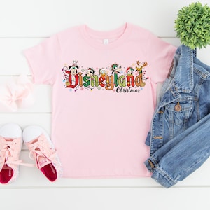 May include: A light pink t-shirt featuring the text "Disneyland Christmas" in colorful lettering with Disney characters. The shirt is paired with a denim jacket, pink sneakers, and a white bow.