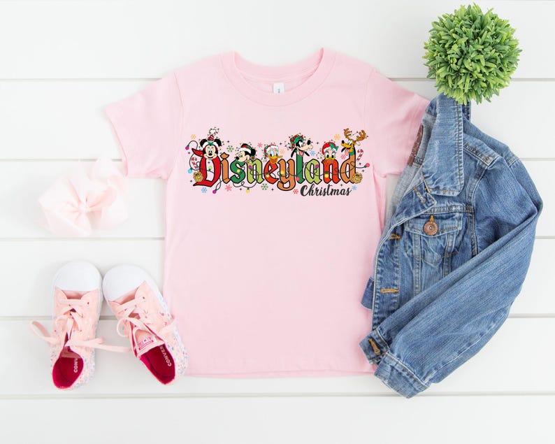 May include: A light pink t-shirt featuring the text "Disneyland Christmas" in colorful lettering with Disney characters. The shirt is paired with a denim jacket, pink sneakers, and a white bow.