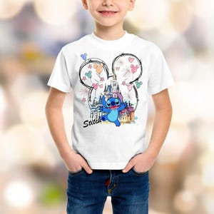 May include: White t-shirt featuring a colorful graphic of the character Stitch from Lilo & Stitch. The design includes a castle, fireworks, and the word "Stitch". The graphic is set within a Mickey Mouse ear outline.