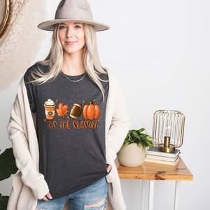 K&ouml;nnte beinhalten: Ein dunkel-melierter grauer T-Shirt mit einem Motiv aus einer Kaffeetasse, einem Blatt, einem Football und einem K&uuml;rbis. Der Text "'Tis the season" ist unter dem Motiv gedruckt.