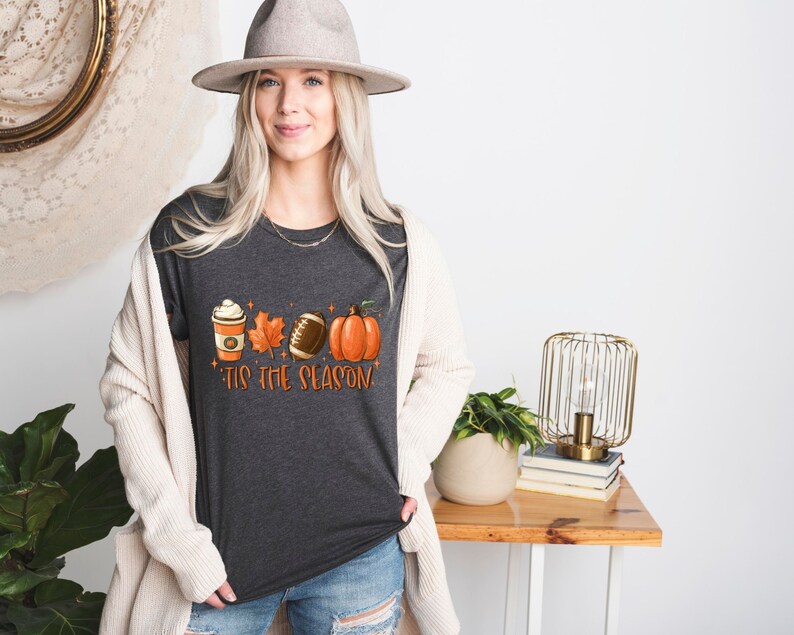 K&ouml;nnte beinhalten: Ein dunkel-melierter grauer T-Shirt mit einem Motiv aus einer Kaffeetasse, einem Blatt, einem Football und einem K&uuml;rbis. Der Text "'Tis the season" ist unter dem Motiv gedruckt.