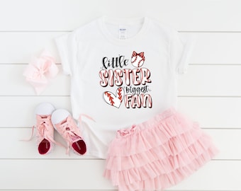 May include: White t-shirt with a red heart and baseball graphic. The text on the shirt reads "little sister biggest fan".
