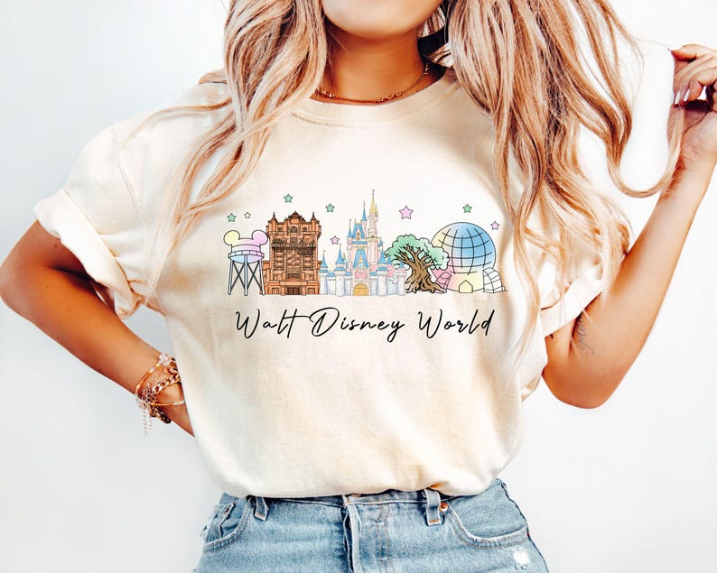 May include: Cream-colored t-shirt featuring a colorful illustration of Walt Disney World landmarks, including the Tower of Terror, Cinderella Castle, and Spaceship Earth. The words "Walt Disney World" are written in a script font below the illustration.