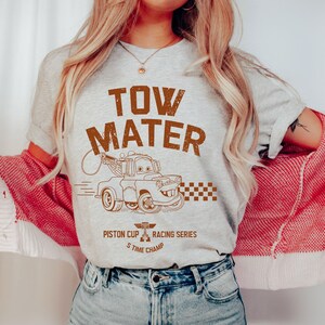 May include: A light gray t-shirt featuring a brown graphic of Tow Mater from the movie Cars. The text "TOW MATER" is above the image, with "Piston Cup Racing Series 5 Time Champ" below. The shirt is paired with blue jeans.