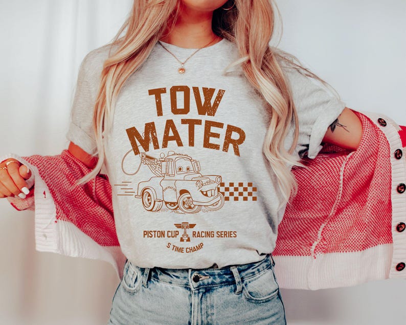 May include: A light gray t-shirt featuring a brown graphic of Tow Mater from the movie Cars. The text "TOW MATER" is above the image, with "Piston Cup Racing Series 5 Time Champ" below. The shirt is paired with blue jeans.
