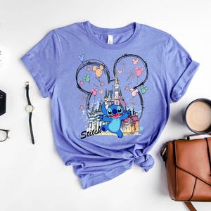 May include: A light purple t-shirt featuring a graphic of Stitch from Lilo & Stitch in front of a castle. The design includes Mickey Mouse ears, fireworks, and the word "Stitch".