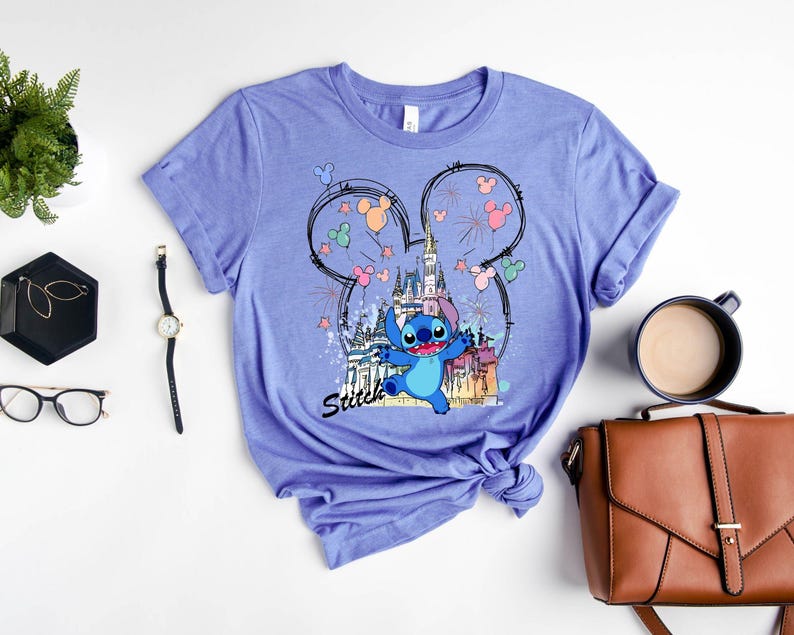 May include: A light purple t-shirt featuring a graphic of Stitch from Lilo & Stitch in front of a castle. The design includes Mickey Mouse ears, fireworks, and the word "Stitch".