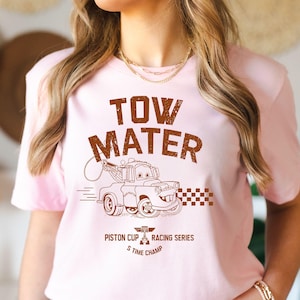 May include: A light pink t-shirt featuring a brown graphic of Tow Mater from the movie Cars. The text "TOW MATER" is above the image, with "Piston Cup Racing Series 5 Time Champ" below. The design includes a checkered flag.