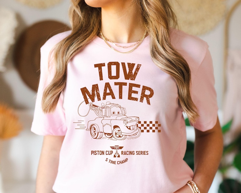 May include: A light pink t-shirt featuring a brown graphic of Tow Mater from the movie Cars. The text "TOW MATER" is above the image, with "Piston Cup Racing Series 5 Time Champ" below. The design includes a checkered flag.