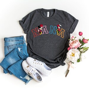 May include: A dark gray t-shirt with a colorful "NANA" graphic featuring Mickey Mouse and Minnie Mouse. The shirt is laid out on a white surface with a pair of white sneakers and blue jeans.