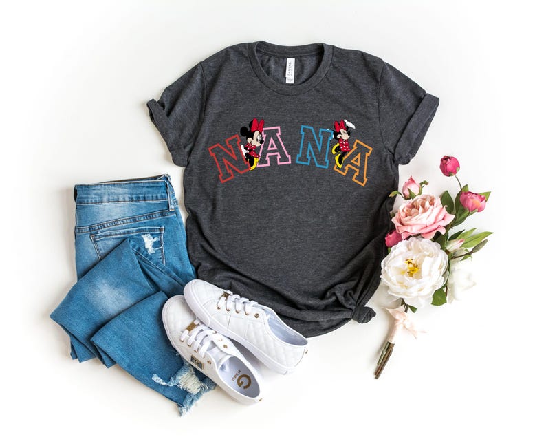 May include: A dark gray t-shirt with a colorful "NANA" graphic featuring Mickey Mouse and Minnie Mouse. The shirt is laid out on a white surface with a pair of white sneakers and blue jeans.