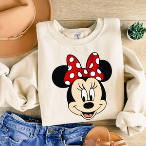 May include: A cream-colored sweatshirt with a cartoon image of Minnie Mouse wearing a red bow with white polka dots.