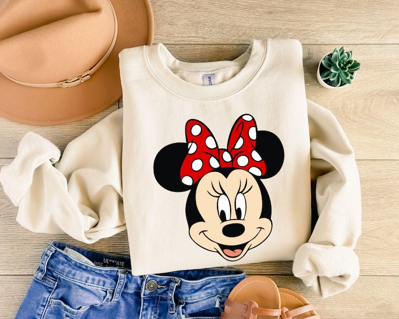 May include: A cream-colored sweatshirt with a cartoon image of Minnie Mouse wearing a red bow with white polka dots.