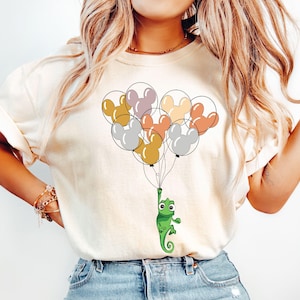 May include: A white t-shirt with a graphic of a chameleon holding onto a bunch of colorful balloons shaped like Mickey Mouse ears.