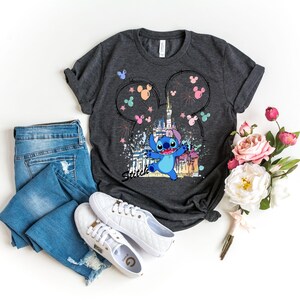 May include: Dark gray t-shirt featuring a colorful graphic of the character Stitch in front of a castle, with Mickey Mouse ears and fireworks. The shirt is paired with blue jeans and white sneakers. A bouquet of pink and white flowers is also present.