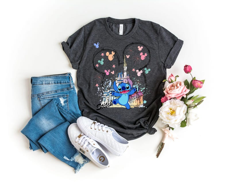 May include: Dark gray t-shirt featuring a colorful graphic of the character Stitch in front of a castle, with Mickey Mouse ears and fireworks. The shirt is paired with blue jeans and white sneakers. A bouquet of pink and white flowers is also present.