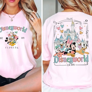 May include: Pale pink t-shirt featuring a Disneyworld design. The front displays Mickey Mouse, Minnie Mouse, Donald Duck, Daisy Duck, and Goofy. The back shows the Disney castle and characters. Text includes "Disneyworld", "Est. 1971", and "Florida".