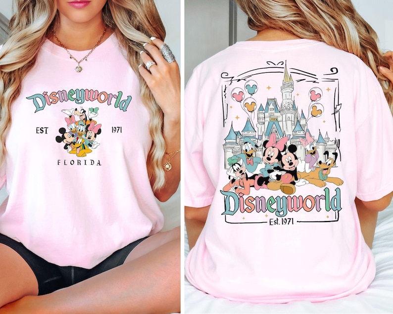 May include: Pale pink t-shirt featuring a Disneyworld design. The front displays Mickey Mouse, Minnie Mouse, Donald Duck, Daisy Duck, and Goofy. The back shows the Disney castle and characters. Text includes "Disneyworld", "Est. 1971", and "Florida".