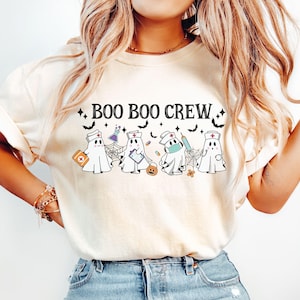 Op de afbeelding: Crèmekleurig T-shirt met de tekst "BOO BOO CREW" boven cartoongeestkarakters verkleed als verpleegsters. De geesten houden medische benodigdheden en een pompoen vast. Zwarte vleermuizen en sterren zijn ook te zien.