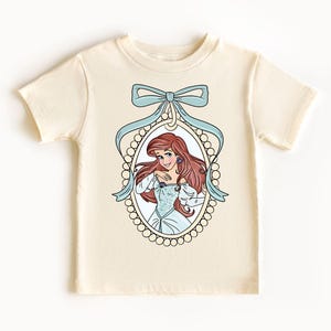 以下が含まれることがあります： クリーム色のTシャツに、真珠で飾られたフレームと青いリボンで囲まれた、リトルマーメイドのアリエルのグラフィックが描かれています。