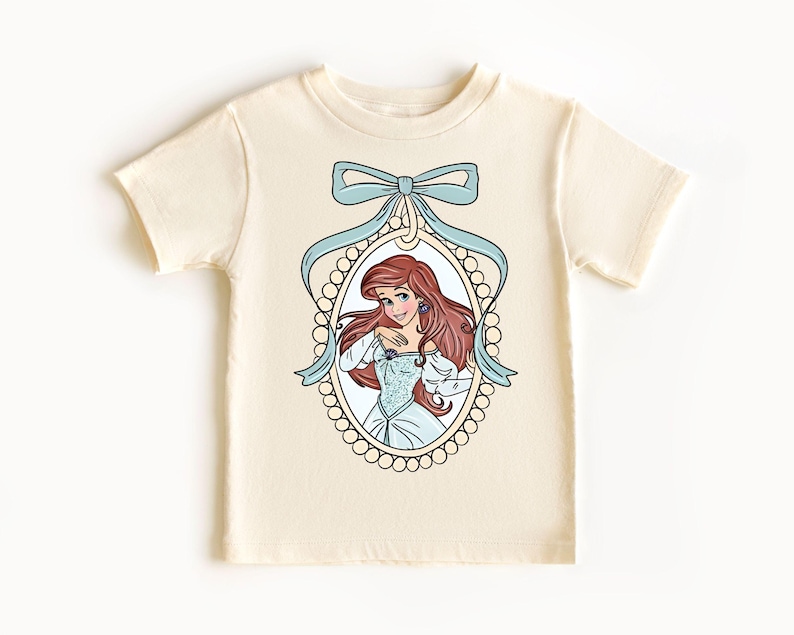 以下が含まれることがあります： クリーム色のTシャツに、真珠で飾られたフレームと青いリボンで囲まれた、リトルマーメイドのアリエルのグラフィックが描かれています。