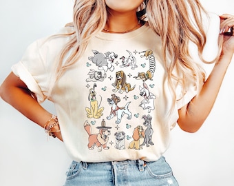 May include: Cream-colored t-shirt featuring a collection of cartoon dog illustrations. The dogs are in various poses and styles, with some wearing hats or accessories. The design includes small blue hearts and cross shapes.