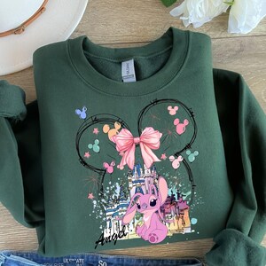 May include: Dark green sweatshirt featuring a graphic of Angel from Lilo & Stitch. The design includes a pink bow, castle, and fireworks, with the word "Angel" in script. The sweatshirt has a crew neck and long sleeves.