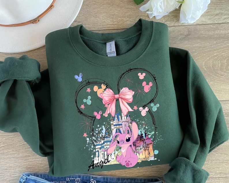 May include: Dark green sweatshirt featuring a graphic of Angel from Lilo & Stitch. The design includes a pink bow, castle, and fireworks, with the word "Angel" in script. The sweatshirt has a crew neck and long sleeves.