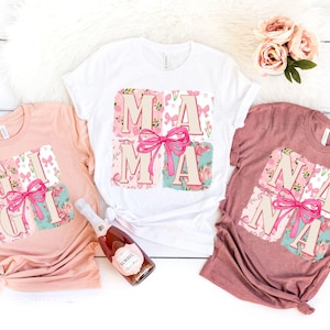 Puede incluir: Tres camisetas en rosa, blanco y malva, cada una con un diseño floral y un lazo. La camiseta blanca dice "MAMA", la rosa dice "GIGI" y la malva dice "NANA". Las camisetas están sobre una superficie blanca.