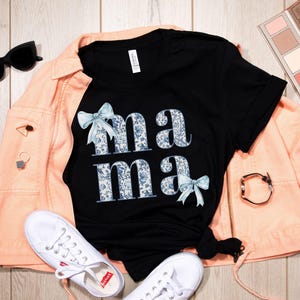 K&ouml;nnte beinhalten: Schwarzes T-Shirt mit einem Blumenmuster und dem Wort "mama" in wei&szlig;en Buchstaben mit blauen Schleifen auf beiden Seiten.