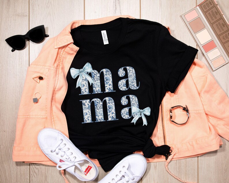 K&ouml;nnte beinhalten: Schwarzes T-Shirt mit einem Blumenmuster und dem Wort "mama" in wei&szlig;en Buchstaben mit blauen Schleifen auf beiden Seiten.