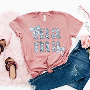 K&ouml;nnte beinhalten: Ein pinkfarbenes T-Shirt mit dem Wort "mama" in blauen und wei&szlig;en Blumenbuchstaben darauf gedruckt. Die Buchstaben sind mit blauen Schleifen verziert. Das Shirt liegt auf einer wei&szlig;en Oberfl&auml;che mit einem Paar rosa Schuhen, einer blauen Jeans und einem Strau&szlig; rosa Blumen.