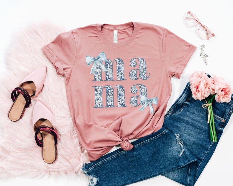 K&ouml;nnte beinhalten: Ein pinkfarbenes T-Shirt mit dem Wort "mama" in blauen und wei&szlig;en Blumenbuchstaben darauf gedruckt. Die Buchstaben sind mit blauen Schleifen verziert. Das Shirt liegt auf einer wei&szlig;en Oberfl&auml;che mit einem Paar rosa Schuhen, einer blauen Jeans und einem Strau&szlig; rosa Blumen.