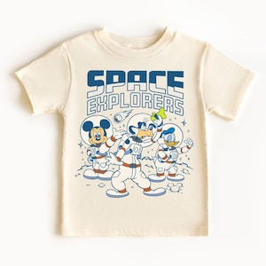 May include: Cream-coloured t-shirt with a graphic of Mickey Mouse, Goofy, and Donald Duck in astronaut suits. The text "SPACE EXPLORERS" is printed above the characters in blue. The design includes a shooting star and stars.