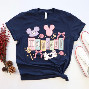May include: Navy blue t-shirt with the word "magical" in pastel colors, featuring Mickey Mouse-themed graphics. Includes a wand, bows, and roses. White sneakers with red trim are placed next to the shirt.