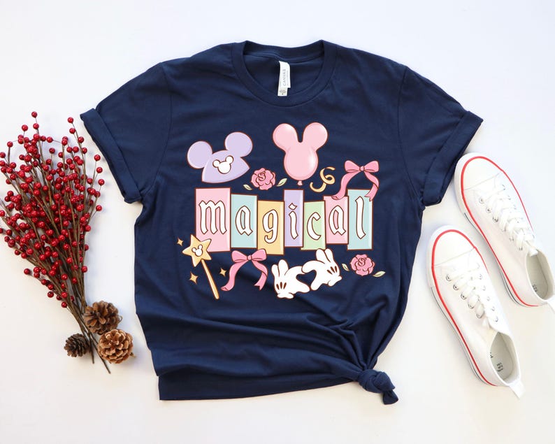 May include: Navy blue t-shirt with the word "magical" in pastel colors, featuring Mickey Mouse-themed graphics. Includes a wand, bows, and roses. White sneakers with red trim are placed next to the shirt.