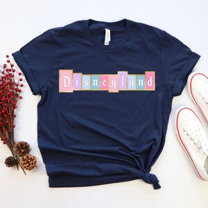 Puede incluir: Camiseta azul marino con la palabra "Disneyland" en colores pastel. El texto est&aacute; en un dise&ntilde;o rectangular. Zapatillas blancas con ribetes rojos est&aacute;n al lado de la camiseta. Un manojo de bayas rojas y pi&ntilde;as est&aacute; a la izquierda.