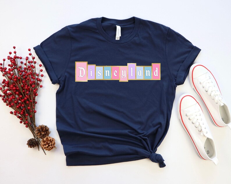Puede incluir: Camiseta azul marino con la palabra "Disneyland" en colores pastel. El texto est&aacute; en un dise&ntilde;o rectangular. Zapatillas blancas con ribetes rojos est&aacute;n al lado de la camiseta. Un manojo de bayas rojas y pi&ntilde;as est&aacute; a la izquierda.