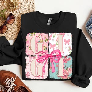 May include: Black crewneck sweatshirt with the word "GIGI" in a pink floral design. The letters are in individual squares with a pink bow in the center. The sweatshirt is surrounded by accessories.