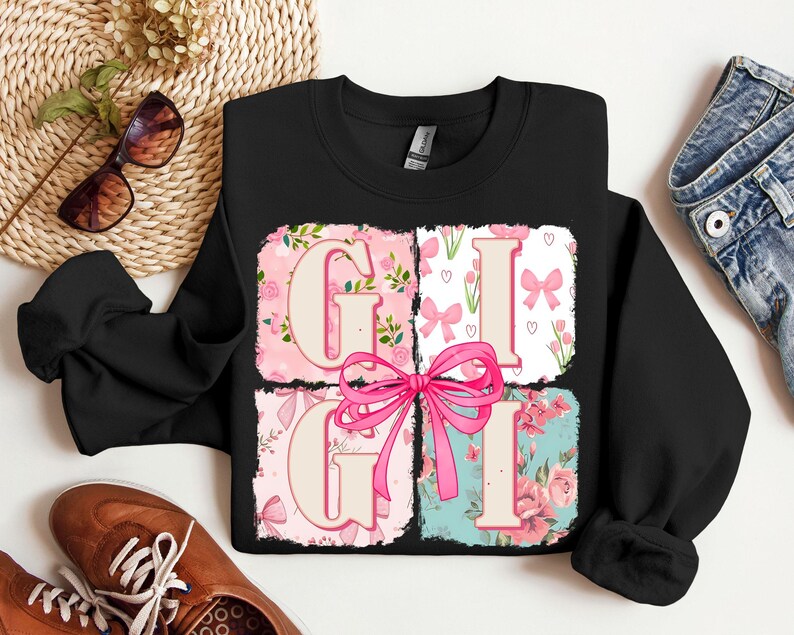 May include: Black crewneck sweatshirt with the word "GIGI" in a pink floral design. The letters are in individual squares with a pink bow in the center. The sweatshirt is surrounded by accessories.