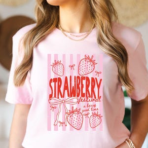 May include: A pale pink t-shirt with a strawberry festival design. The graphic includes red text and drawings of strawberries and bows set against a pink and white striped backdrop. The text reads "STRAWBERRY festival a berry good time."