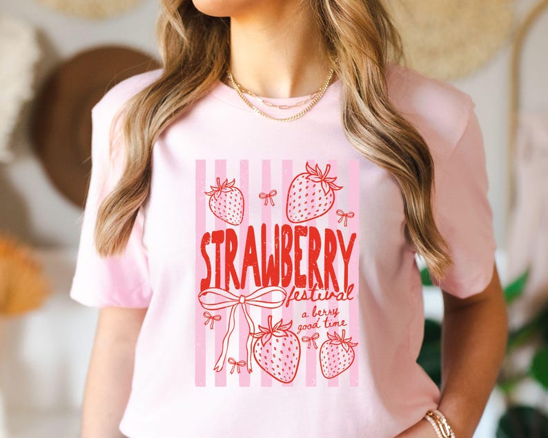 May include: A pale pink t-shirt with a strawberry festival design. The graphic includes red text and drawings of strawberries and bows set against a pink and white striped backdrop. The text reads "STRAWBERRY festival a berry good time."