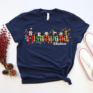 May include: Navy blue t-shirt featuring the text "Disneyland Christmas" in colorful lettering, with Disney characters. The shirt is paired with white sneakers with red trim. A sprig of red berries and pinecones are on the left.
