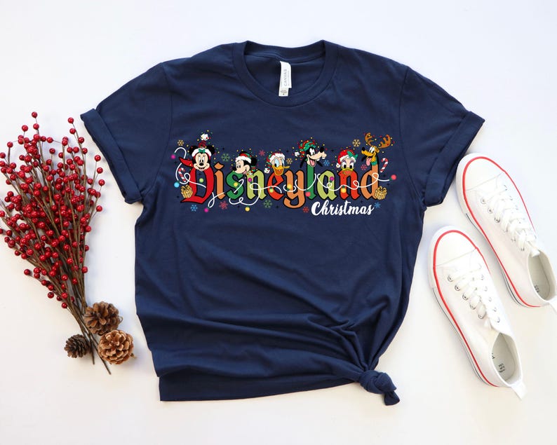 May include: Navy blue t-shirt featuring the text "Disneyland Christmas" in colorful lettering, with Disney characters. The shirt is paired with white sneakers with red trim. A sprig of red berries and pinecones are on the left.