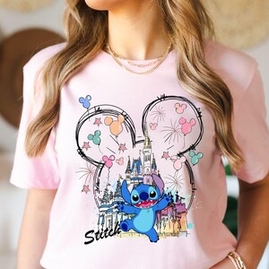 May include: A light pink t-shirt featuring a cartoon graphic of Stitch from Lilo & Stitch. The design includes a castle, fireworks, and Mickey Mouse ears with balloons. The word "Stitch" is written in black script below the character.