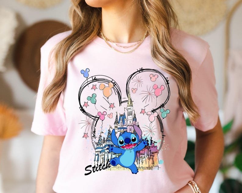 May include: A light pink t-shirt featuring a cartoon graphic of Stitch from Lilo & Stitch. The design includes a castle, fireworks, and Mickey Mouse ears with balloons. The word "Stitch" is written in black script below the character.