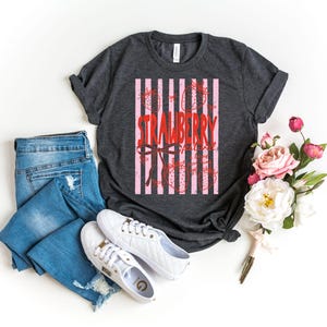 May include: A charcoal grey t-shirt with a pink and white striped design featuring the word "STRAWBERRY" in red script. The shirt is paired with blue jeans and white trainers. A bouquet of pink and white flowers is also present.