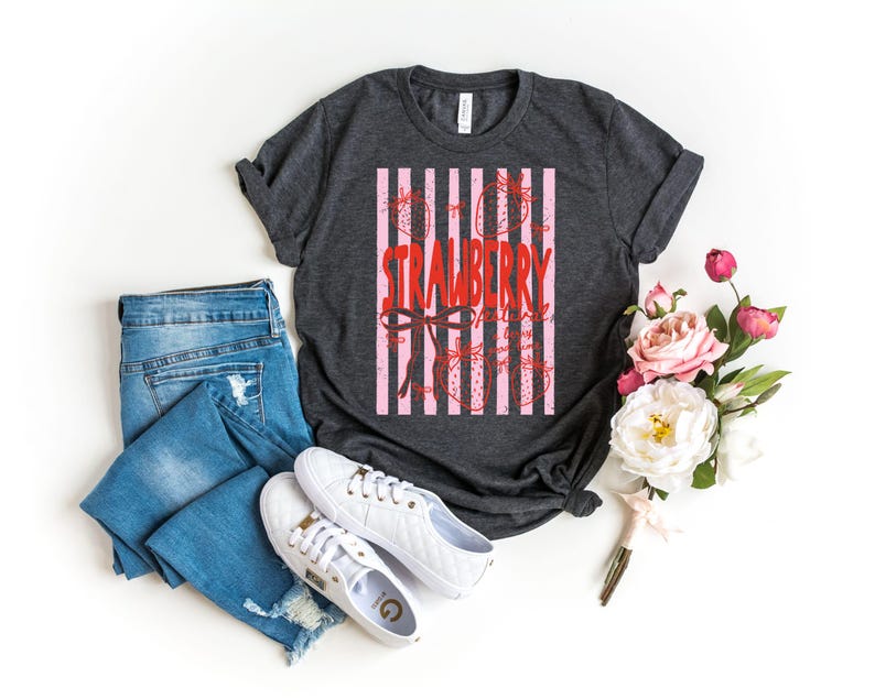 May include: A charcoal grey t-shirt with a pink and white striped design featuring the word "STRAWBERRY" in red script. The shirt is paired with blue jeans and white trainers. A bouquet of pink and white flowers is also present.