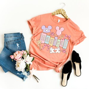 May include: A peach-colored t-shirt with the word "magical" in pastel colors, featuring Mickey Mouse-inspired designs and other graphics. The shirt is paired with blue jeans, black flats, and a bouquet of flowers. The shirt is hanging on a gold hanger.