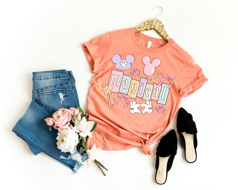 May include: A peach-colored t-shirt with the word "magical" in pastel colors, featuring Mickey Mouse-inspired designs and other graphics. The shirt is paired with blue jeans, black flats, and a bouquet of flowers. The shirt is hanging on a gold hanger.